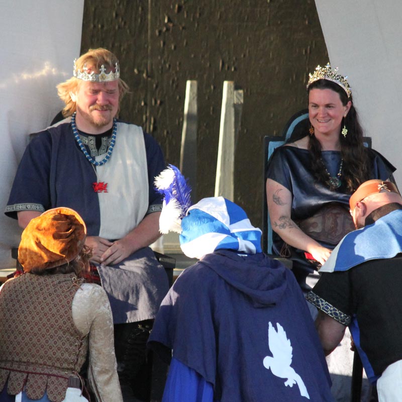 King and queen wearing white and blue garb sit on thrones while people bow